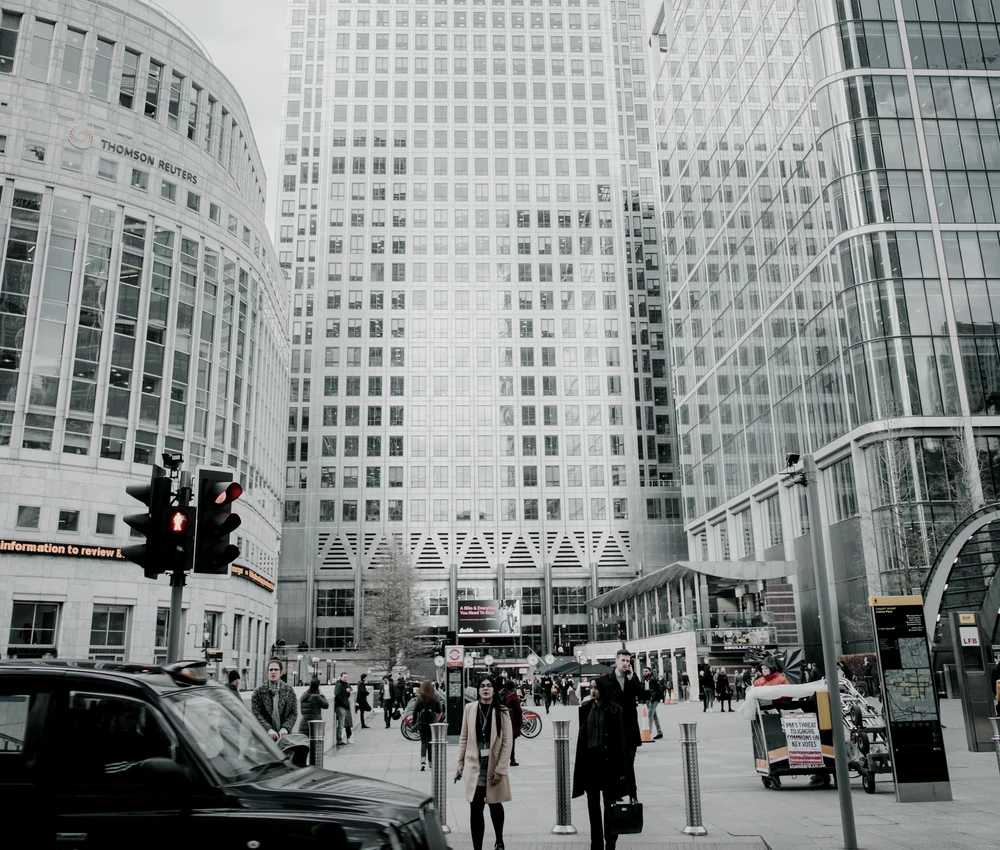 Pedestrians and street traffic beneath glass office towers in Canary Wharf, London, rendered in restrained steel-grey tones