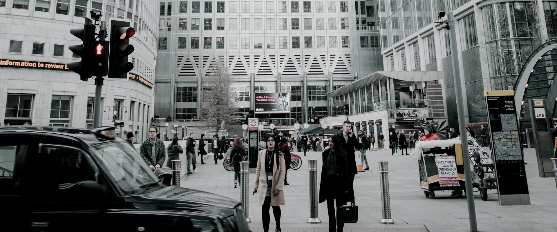 Pedestrians and street traffic beneath glass office towers in Canary Wharf, London, rendered in restrained steel-grey tones